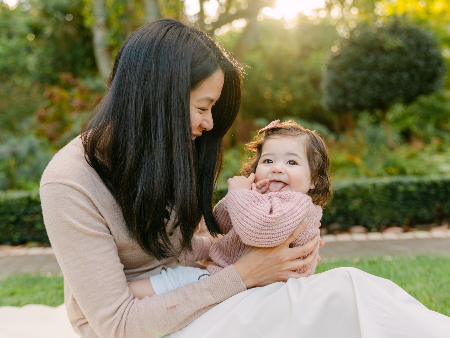 A mother smiles lovingly at her baby girl in a pink sweater during an outdoor family photo session.