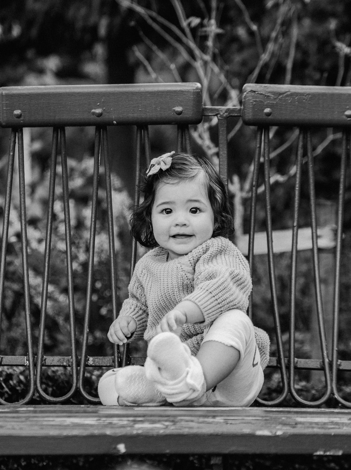 Black and white photo of an adorable toddler girl sitting on a bench outdoors, wearing a knit sweater with a flower in her hair.