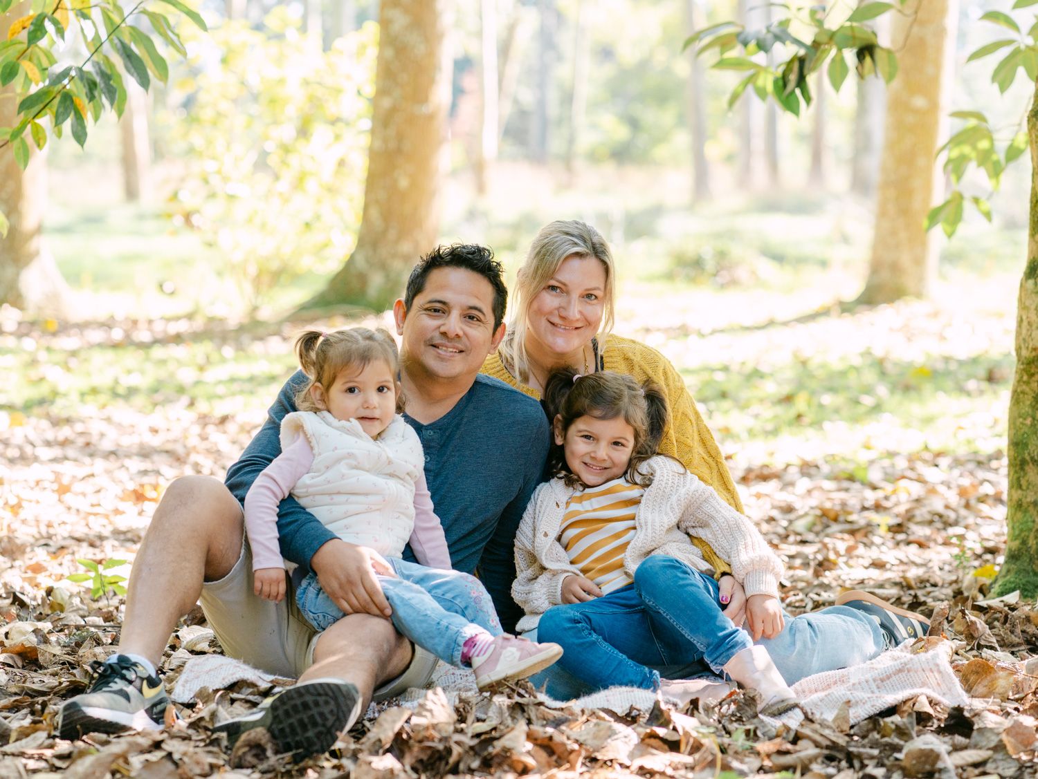 Happy family of four sitting among fallen leaves in a sunny park, smiling together outdoors.