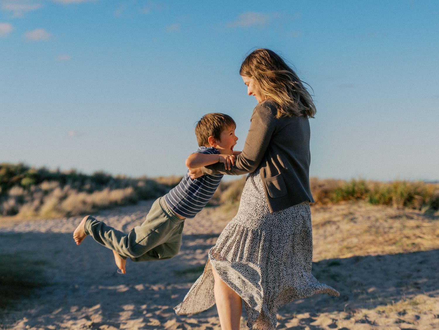 Woman swinging a young boy on a sandy beach under a blue sky, sharing a joyful moment together.