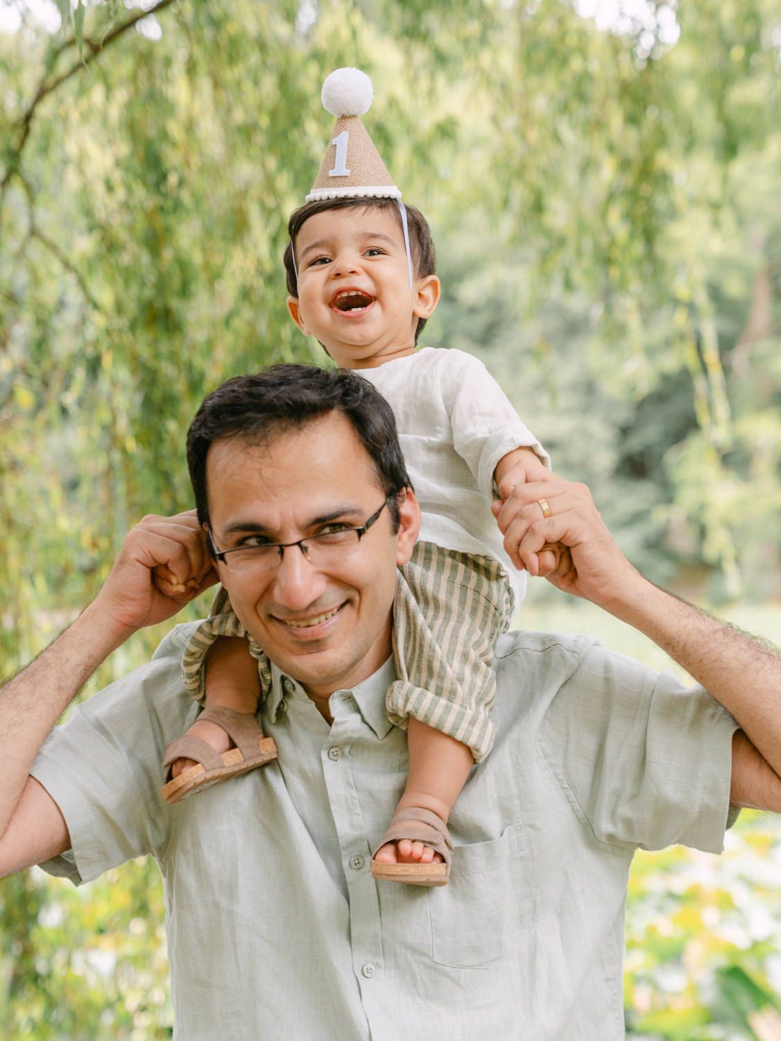 A laughing toddler wearing a birthday hat sits on a smiling man's shoulders outdoors among green trees.
