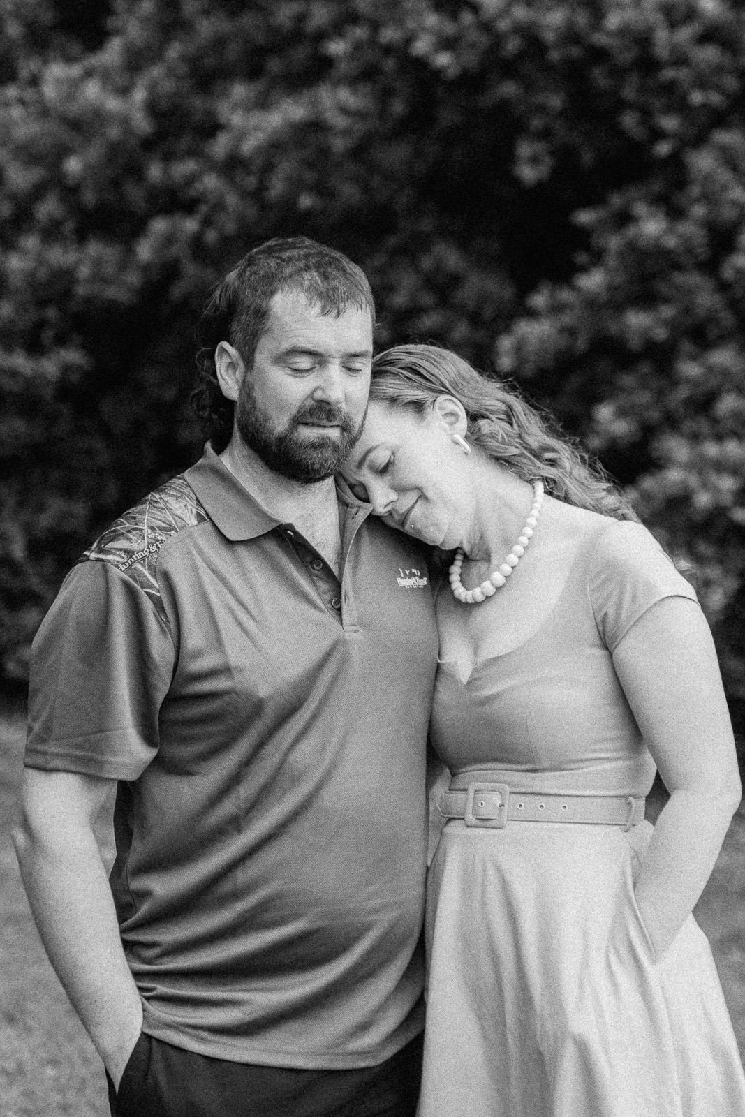 Couple shares tender moment outdoors, woman resting head on mans shoulder in black and white photo.
