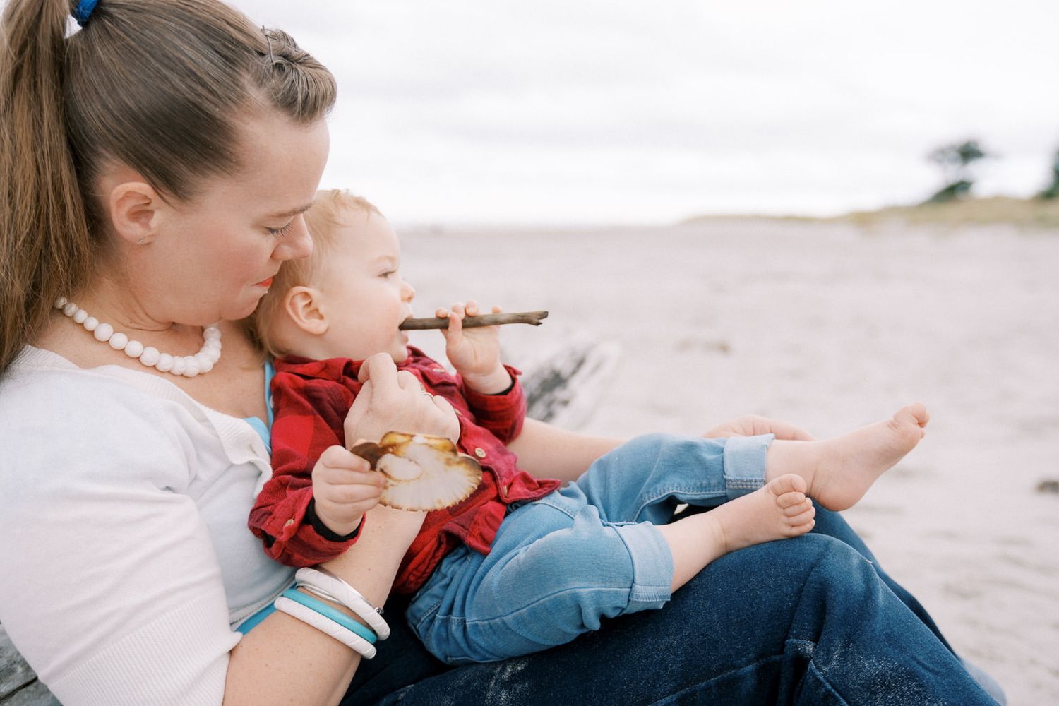 Mother kissing baby on beach, infant in red plaid shirt holding stick while sitting on mom's lap.