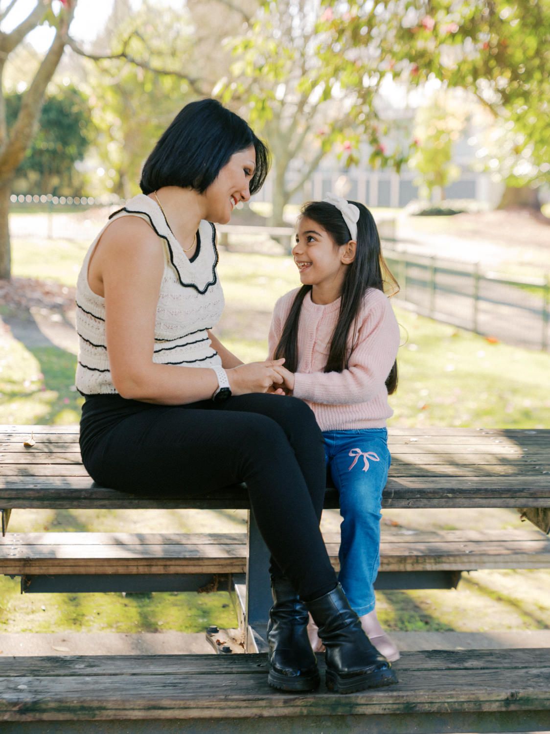 A woman and young girl sit on a park bench smiling at each other, sharing a warm moment outdoors.