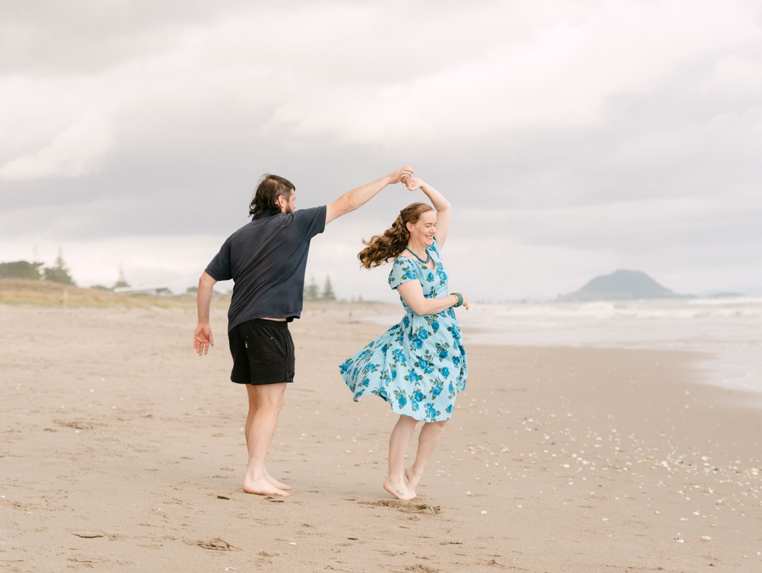 Couple dancing on a beach, woman in blue floral dress being twirled by man in dark shirt and black shorts.