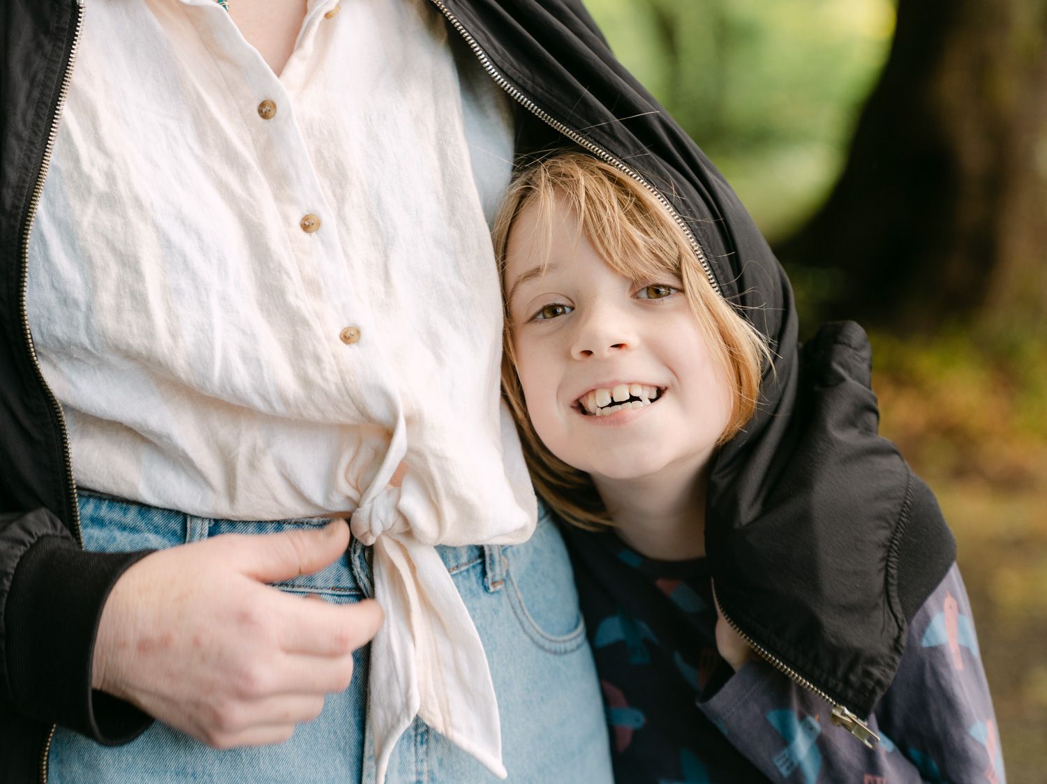 A smiling child with blonde hair leans against an adult outdoors, wearing a black hoodie among green trees.