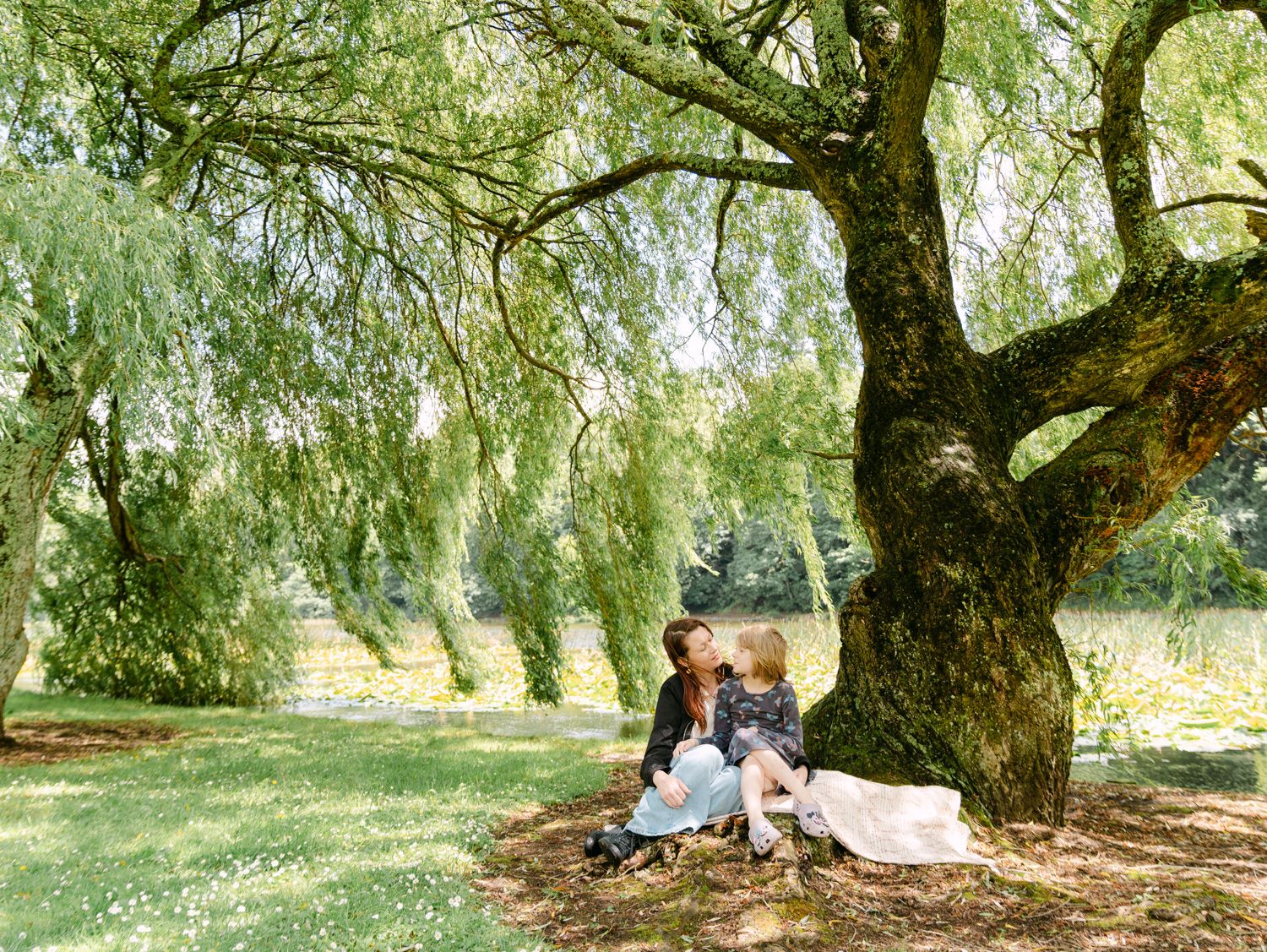 Mother and child sitting together under a large willow tree in a lush green park on a sunny day.