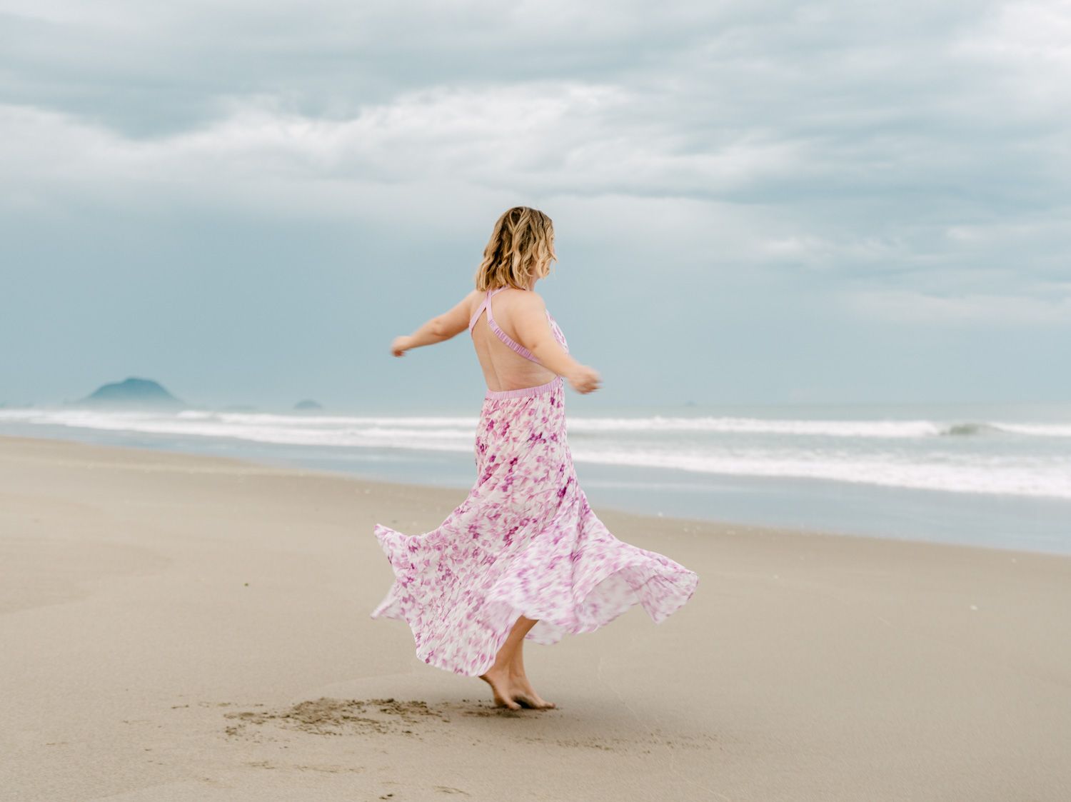 Woman in floral pink maxi dress twirling on sandy beach with ocean waves and cloudy sky backdrop.