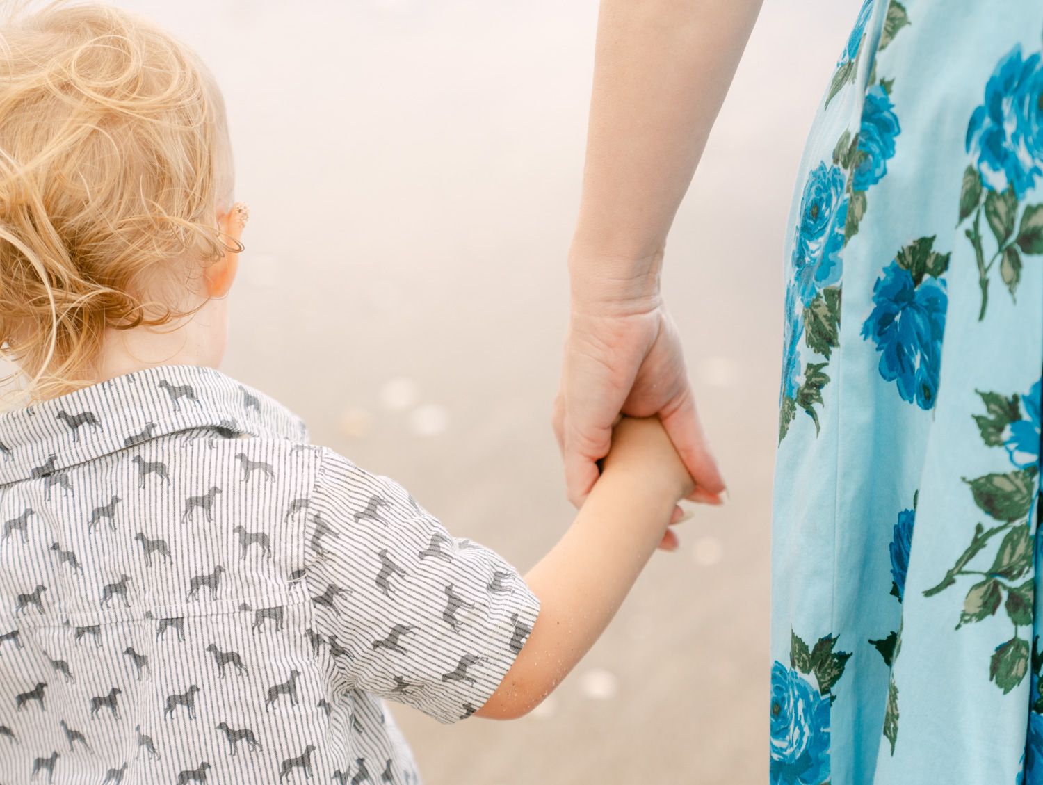 Close-up of a mother holding her toddler's hand at the beach, wearing a floral blue dress.