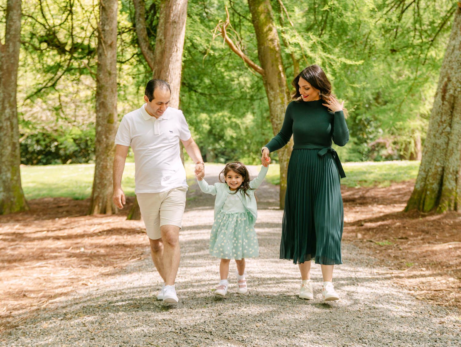 Family of three walking hand-in-hand on a tree-lined path, parents swinging young girl between them.