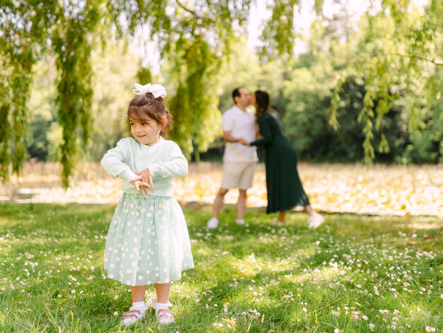 A toddler girl in a white dress plays in a sunny park while her parents embrace under willow trees.