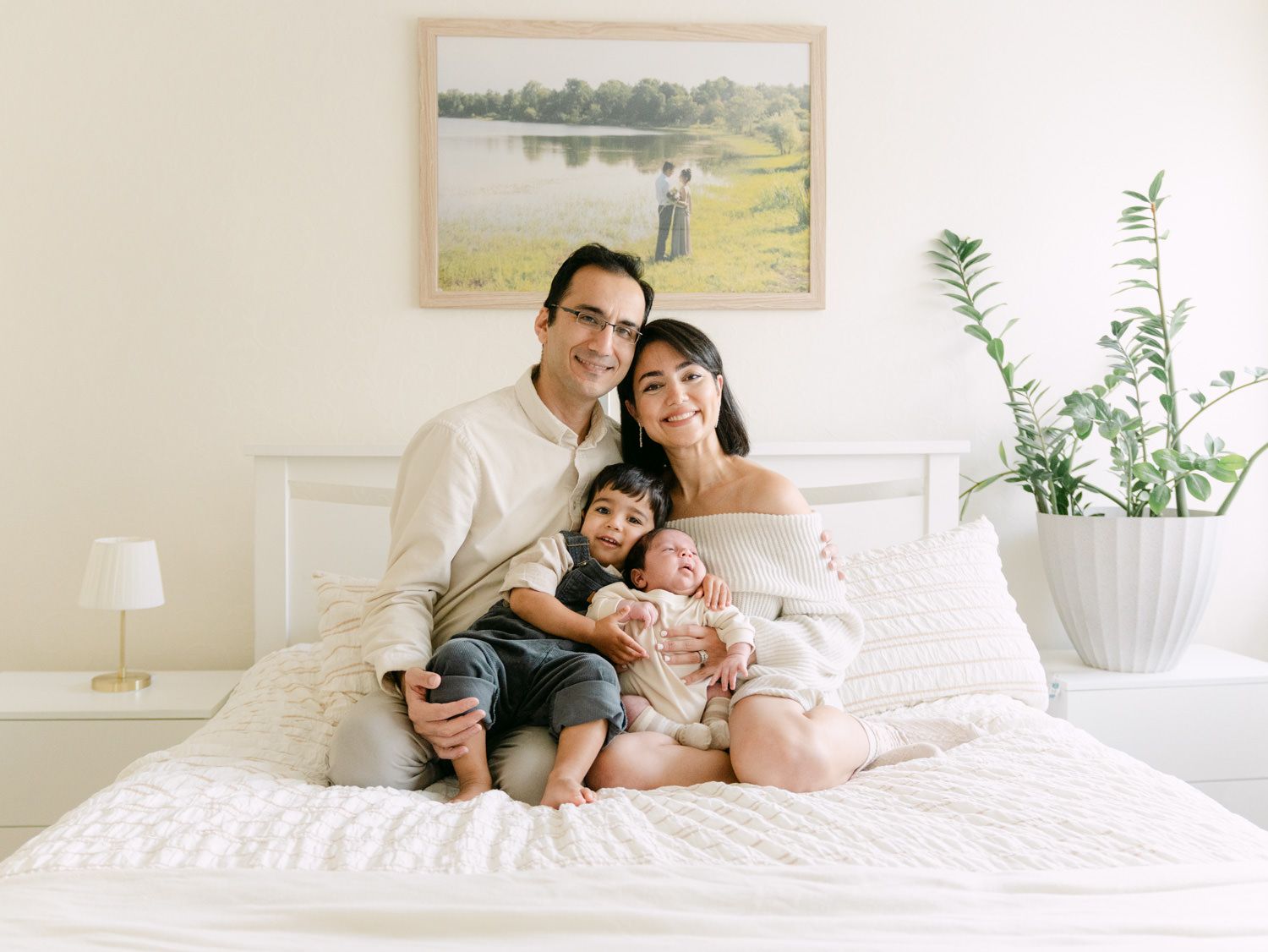 Happy family of four posing on a white bed in a bright bedroom with green plants and artwork on the wall.