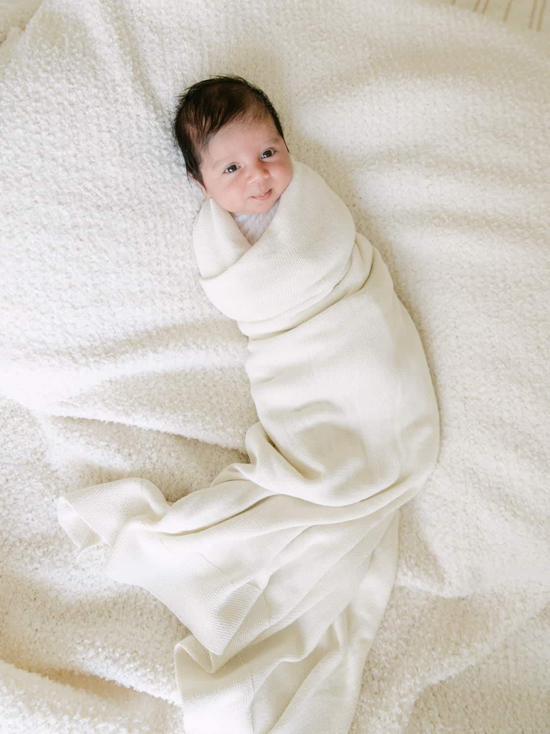 Newborn baby swaddled in a soft ivory blanket, lying on a white textured surface, looking up with a gentle smile.