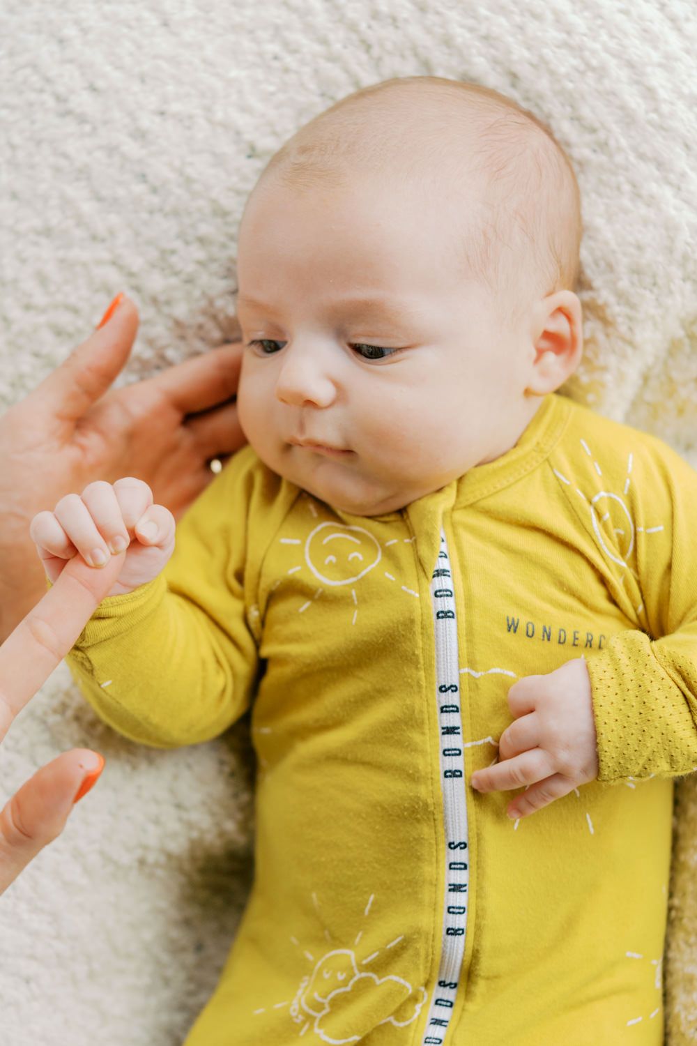 Newborn baby in yellow Bonds wondersuit onesie lying on white blanket, grasping adult finger.