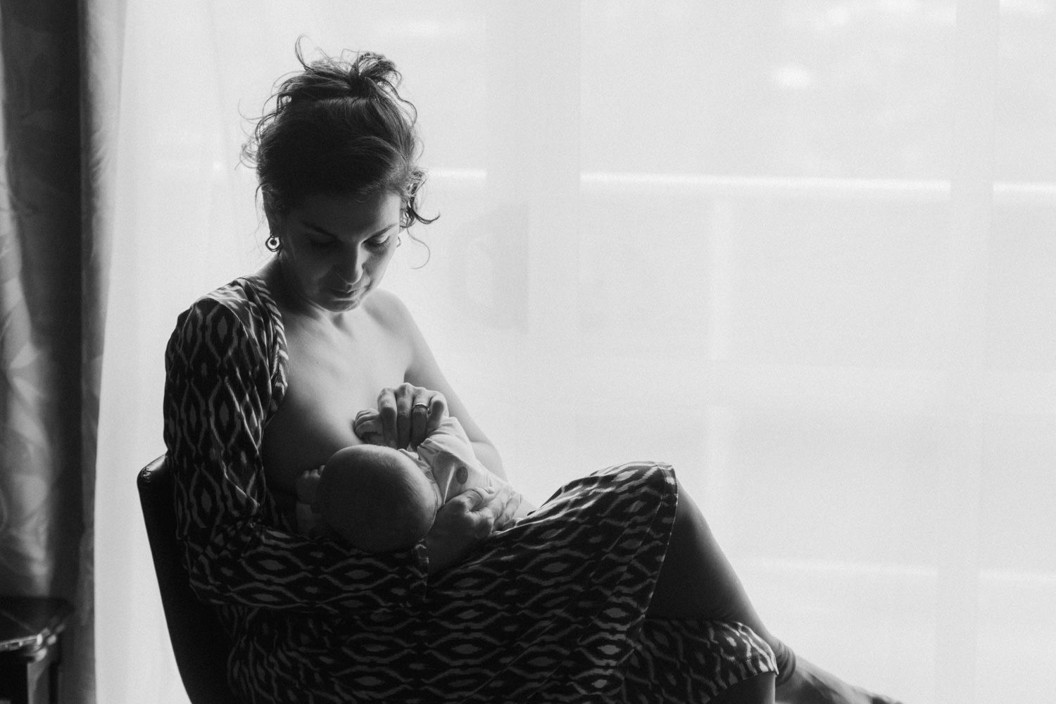 Black and white photo of a mother breastfeeding her newborn baby while seated in a chair near a window.