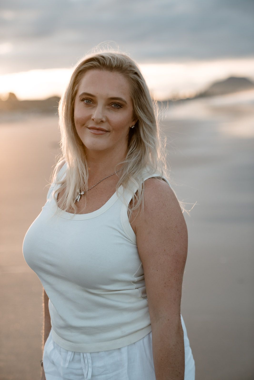 Blonde woman in white tank top smiling on a beach at sunset with cloudy sky background.
