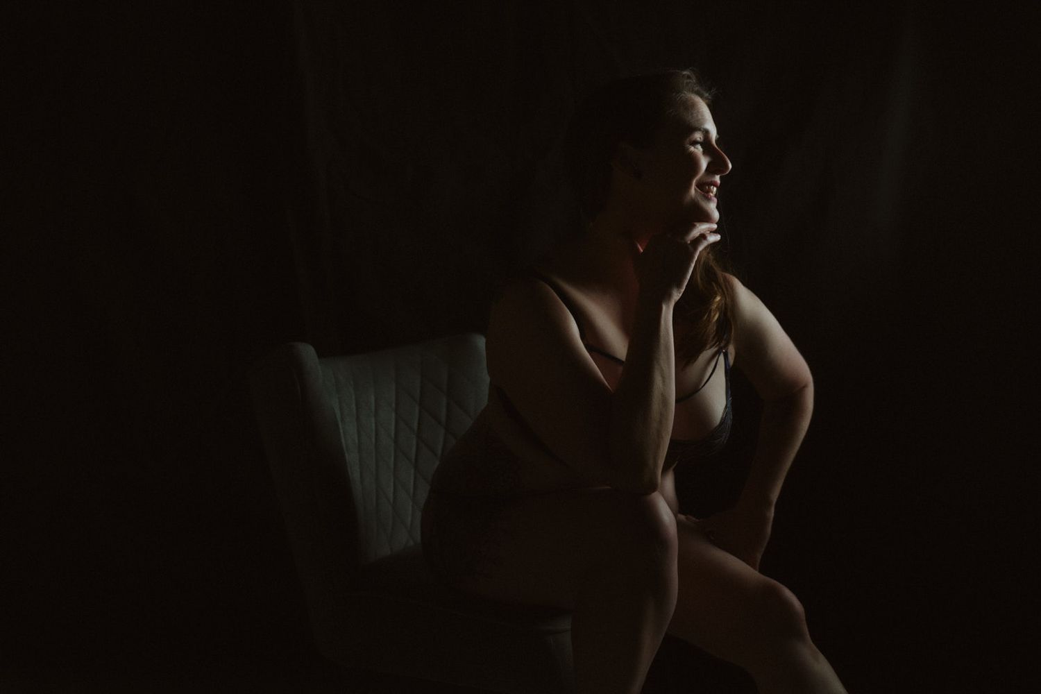 Woman in lingerie sitting pensively in dramatic low-key lighting, contemplative boudoir portrait photography.