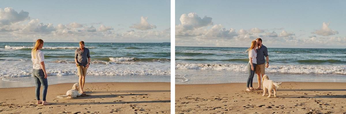 Couple with a white dog standing on a sandy beach with ocean waves and cloudy sky in the background.
