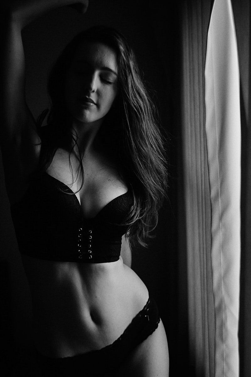 Black and white boudoir photo of a woman in lingerie posing gracefully near a window with soft lighting.