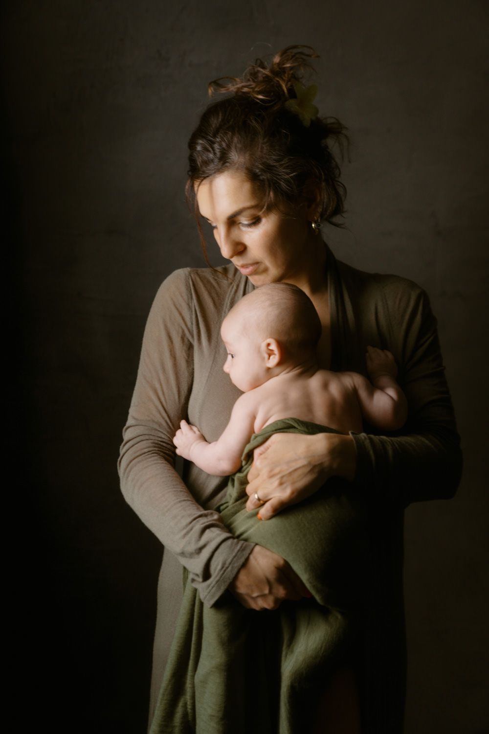 A mother in an olive dress tenderly holds her newborn baby wrapped in a green blanket against a dark background.
