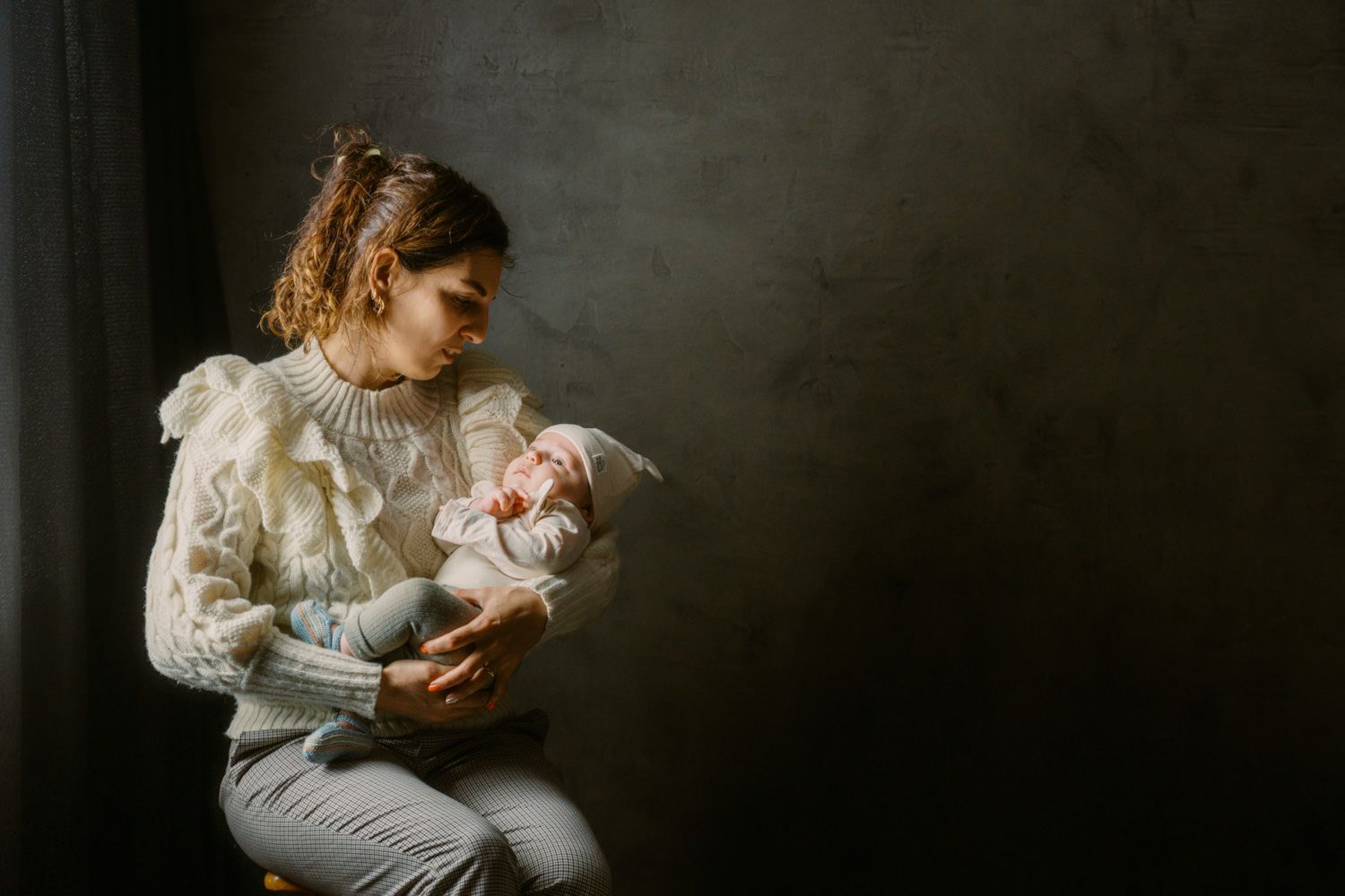 A mother tenderly cradles her newborn baby while seated near a window in soft, natural light.