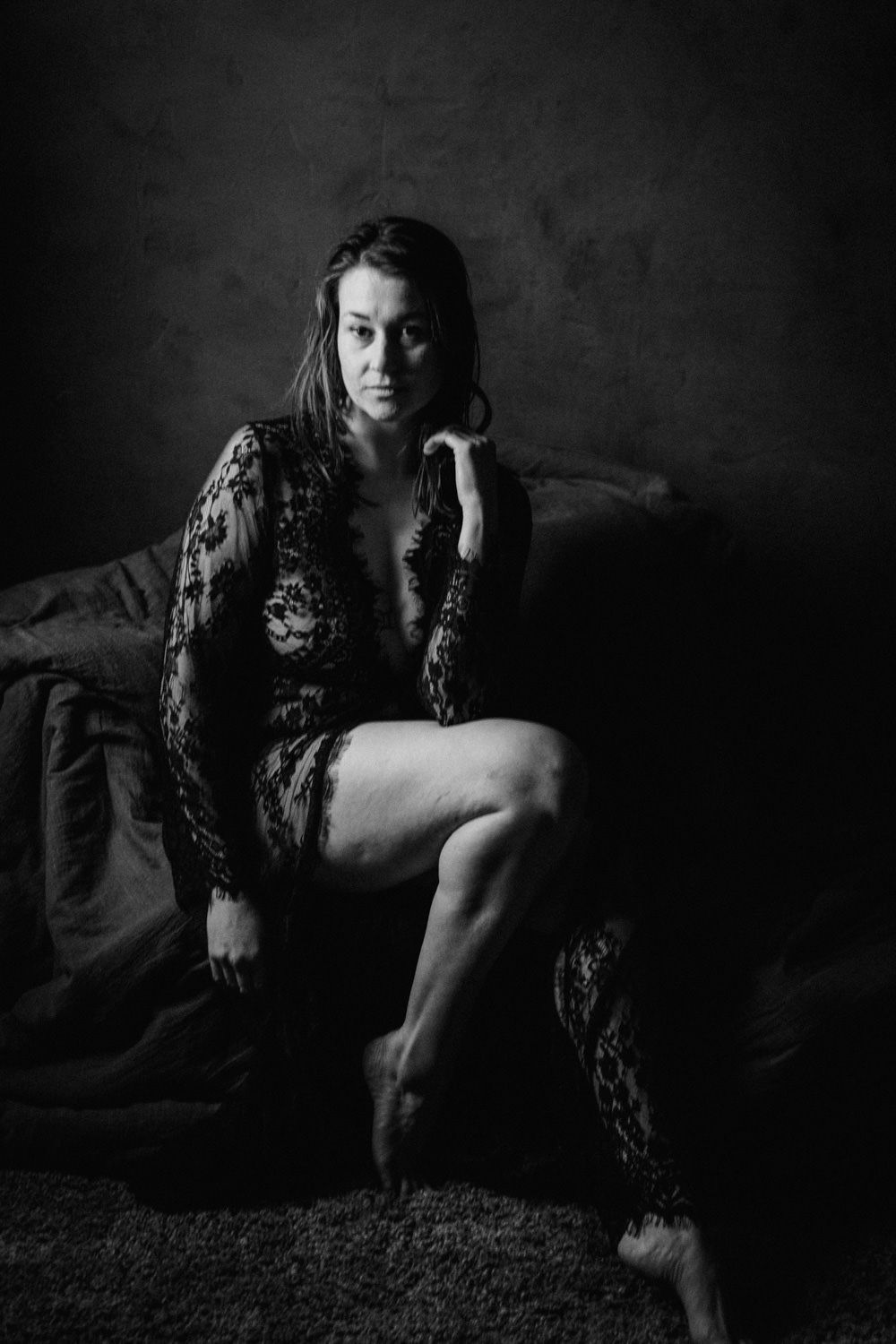 Black and white boudoir photo of a woman in lace bodysuit sitting on a bed in dramatic moody lighting.