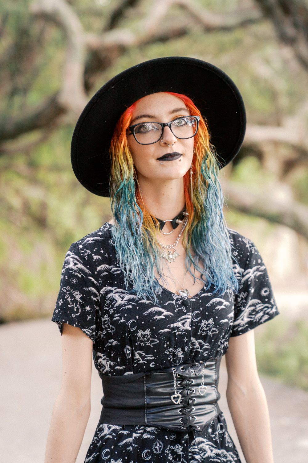 Young woman with rainbow hair wearing black hat, glasses, dark lipstick, and gothic corset outfit outdoors.