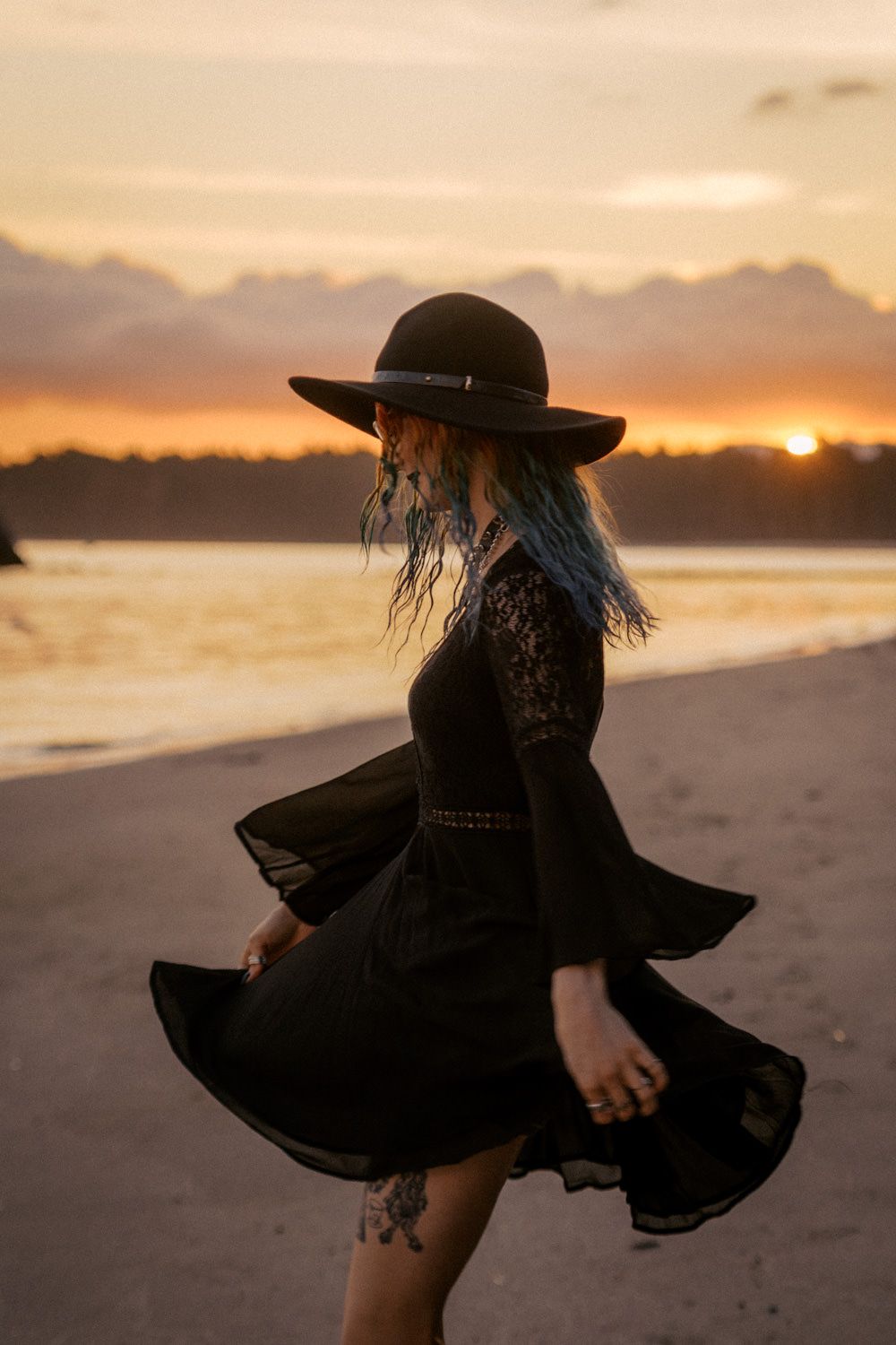Woman in black lace dress and wide-brim hat twirling on beach at golden sunset with blue-dyed hair.