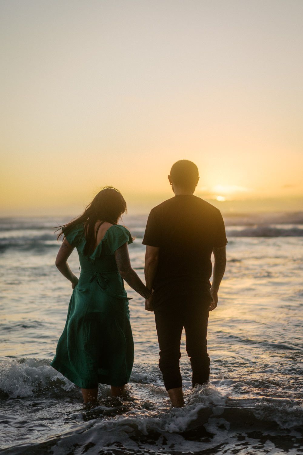 Couple holding hands on beach at sunset, woman in green dress and man in black shirt standing in ocean waves.