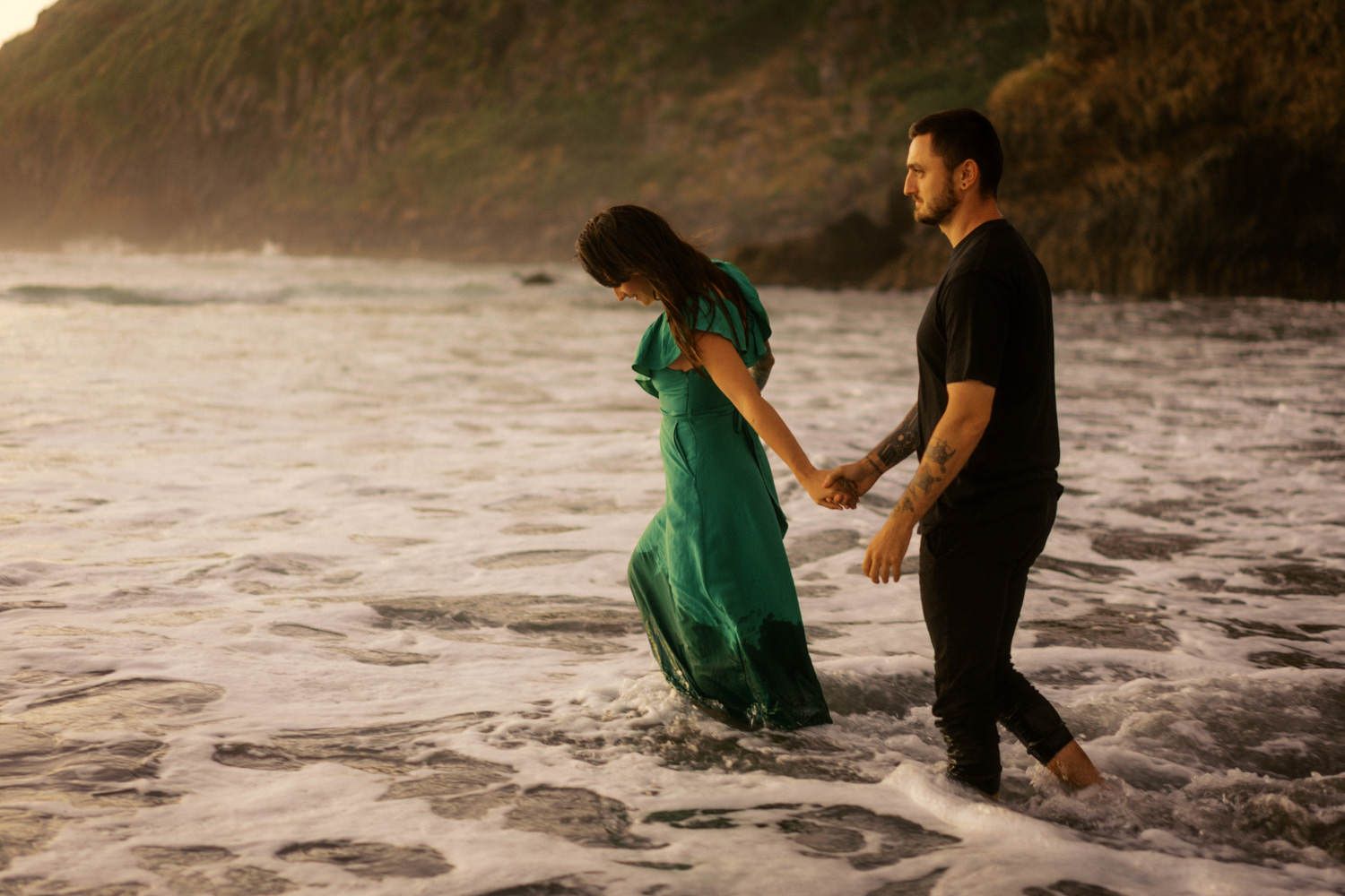 Couple holding hands walking through ocean waves at sunset, woman in flowing green dress with man in black.