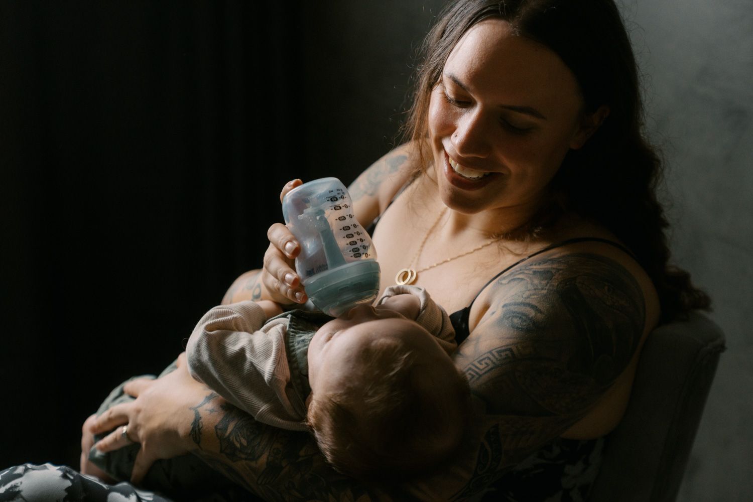 Smiling tattooed mother bottle feeding her newborn baby while sitting in a chair in soft natural light.