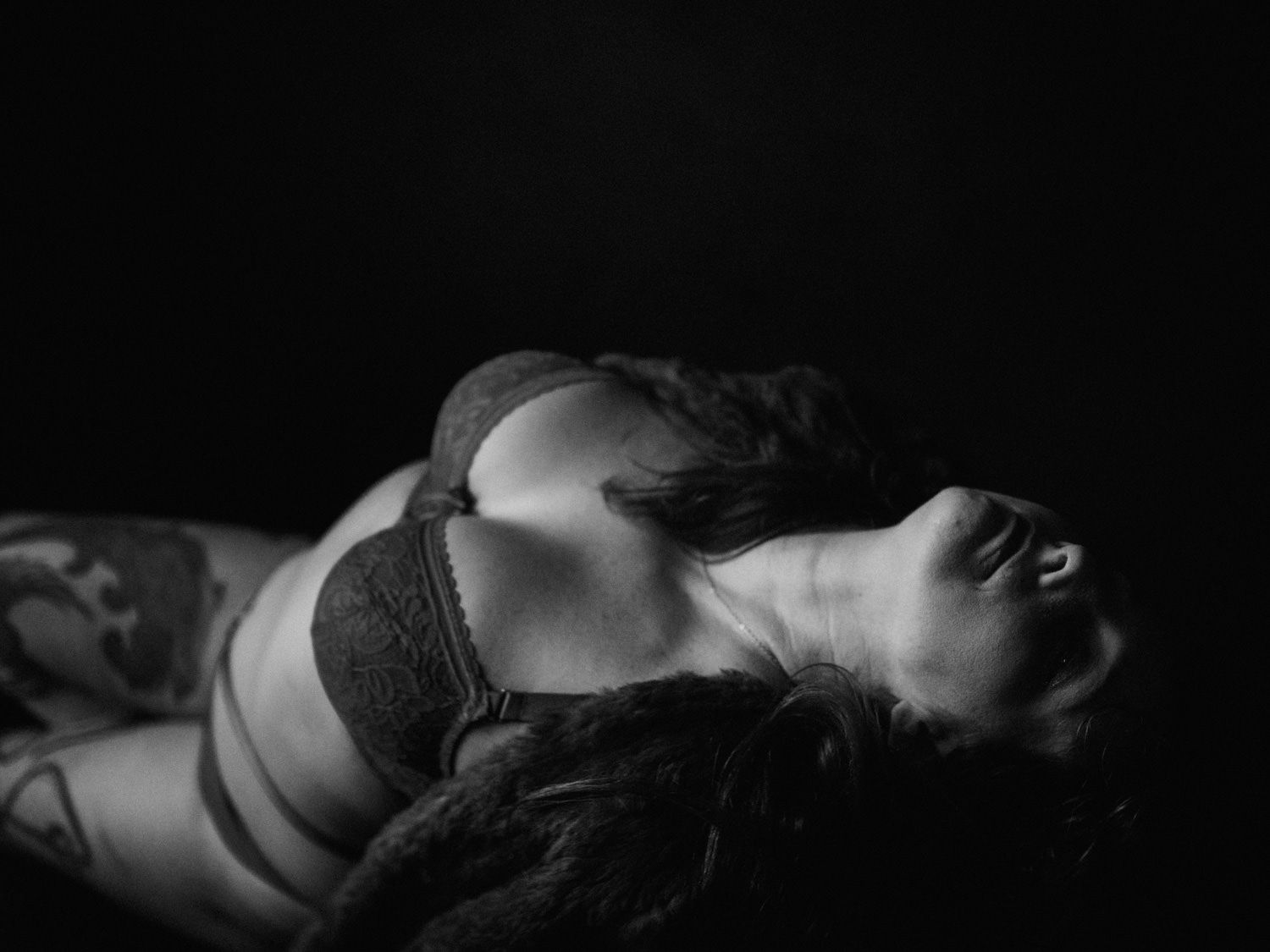 Black and white boudoir photo of a tattooed woman in lace lingerie posing dramatically against a dark background.
