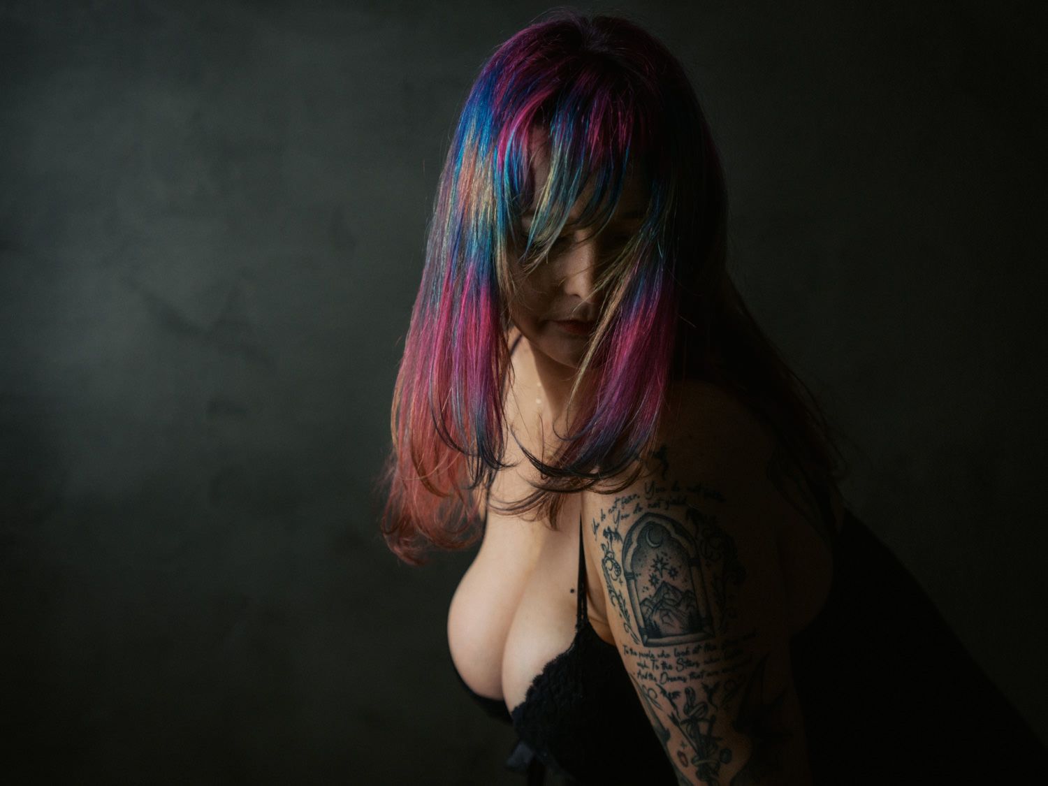 Woman with vibrant multicolored hair and tattoos posing against a dark background.