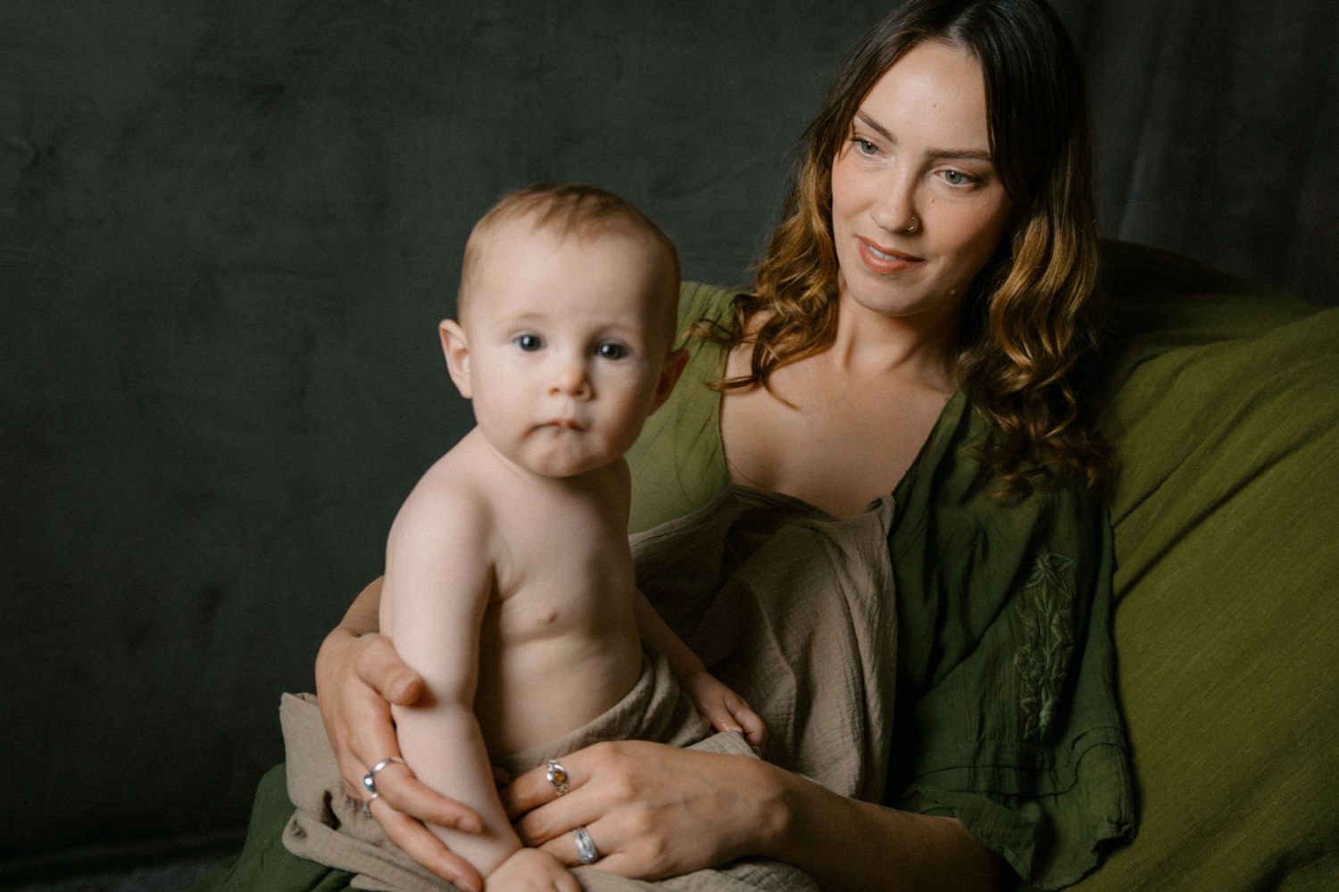 A young mother in a green dress tenderly holds her shirtless baby boy while sitting on a green chair.