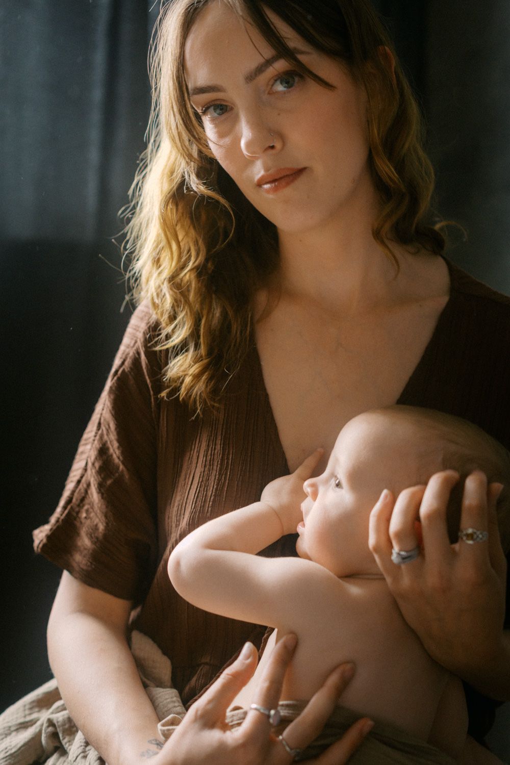 A young mother with wavy hair holds her infant baby in her arms against a dark background.