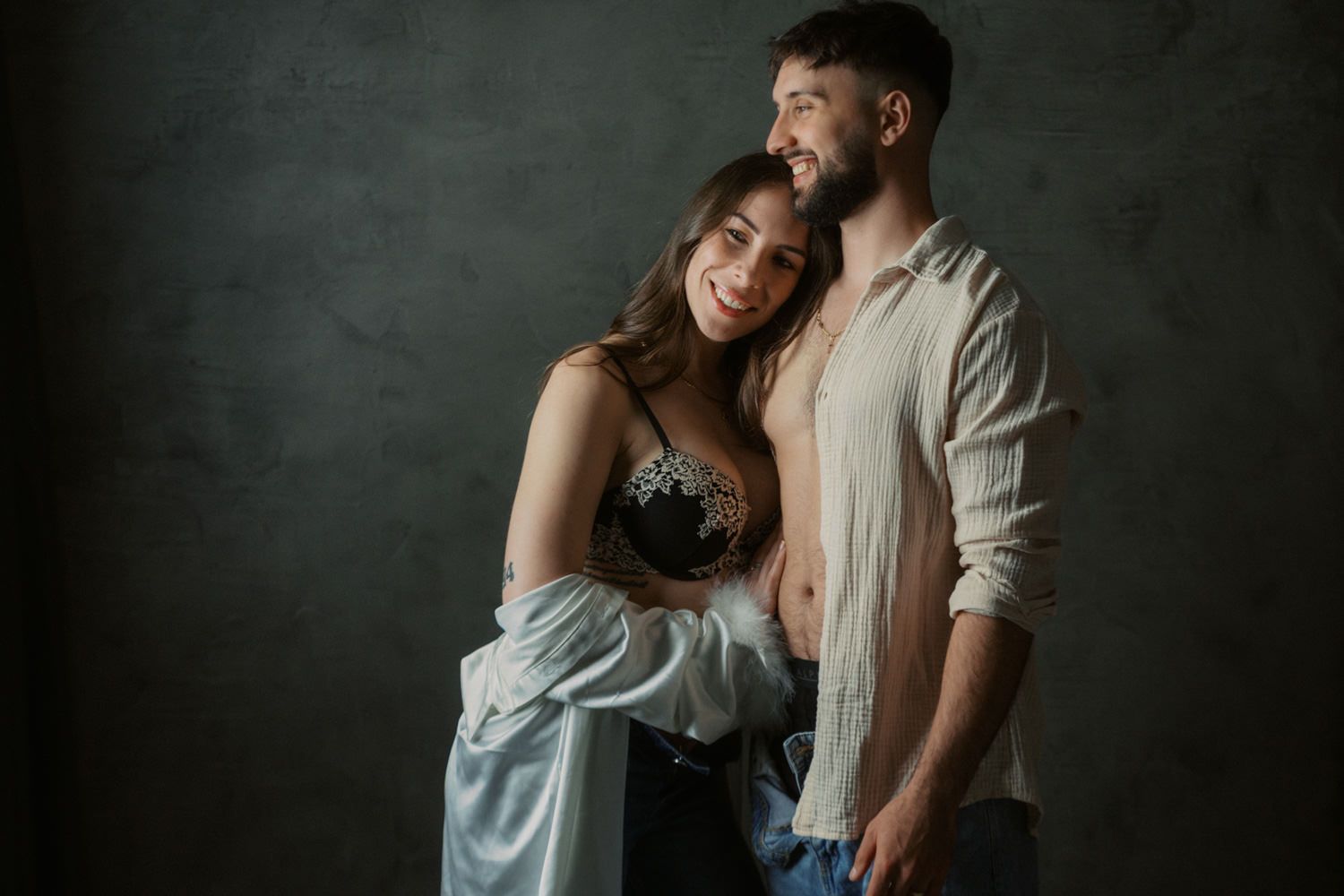 A stylish couple poses intimately in a studio, woman in black lace bra and blue shirt, man in open white shirt.