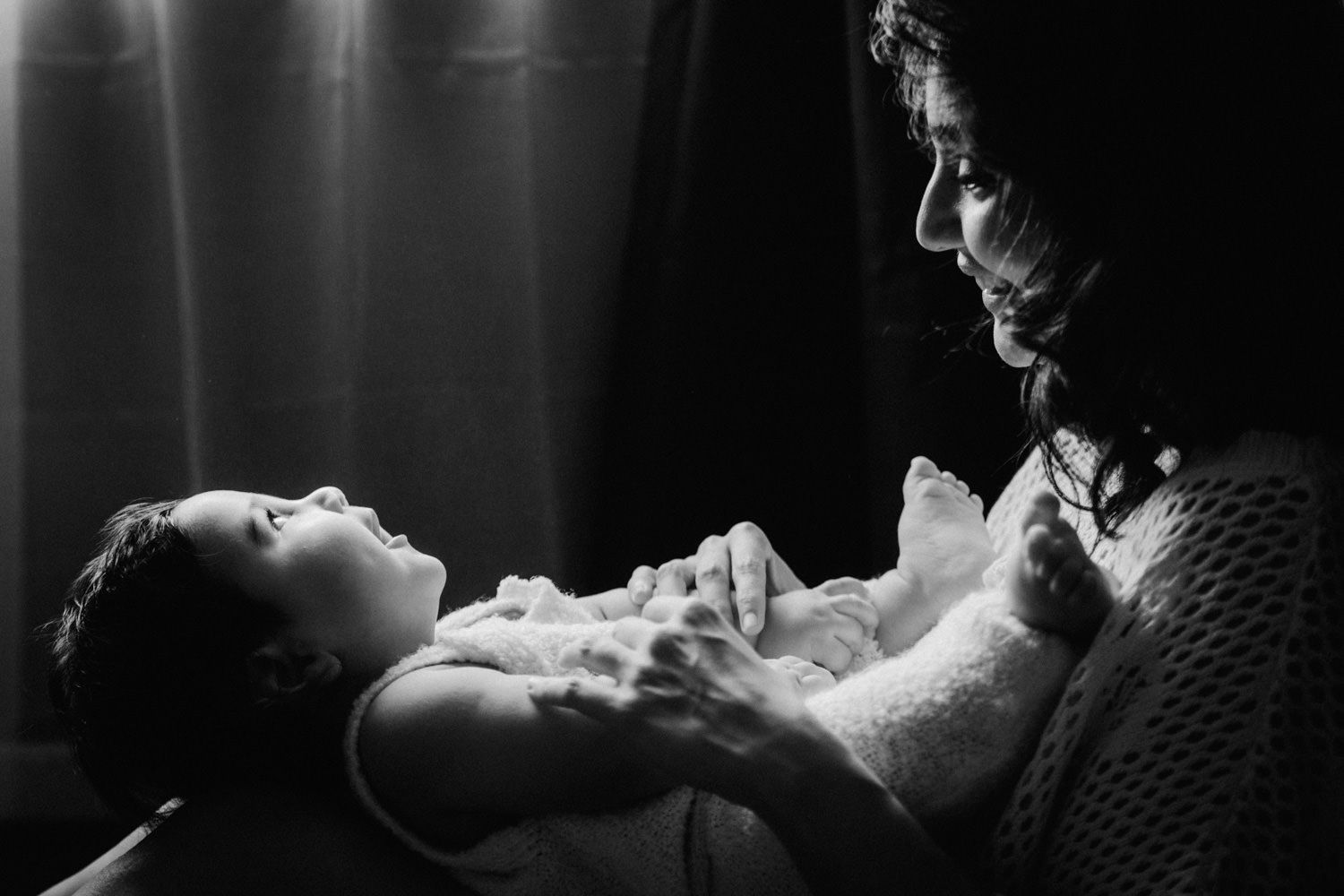 Black and white photo of a mother tenderly cradling her newborn baby in soft, dramatic lighting.