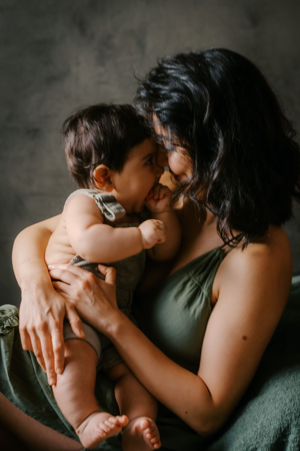 A mother tenderly holds her baby close, touching foreheads in an intimate moment against a dark studio backdrop.