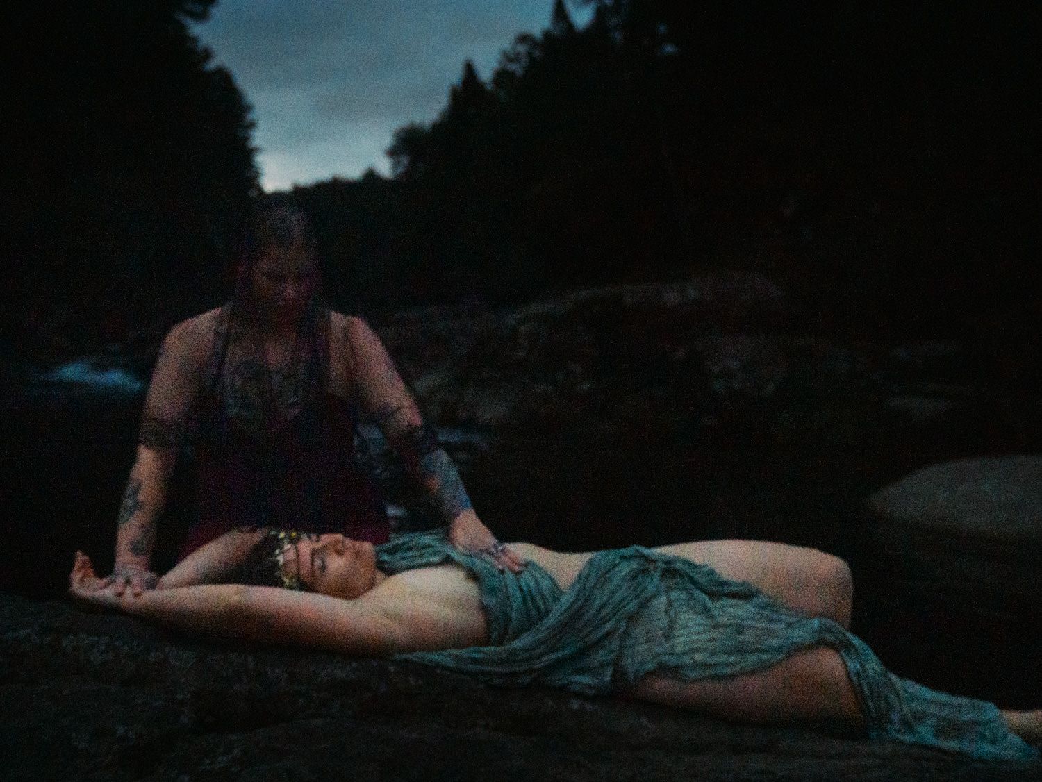 Two women in a dramatic outdoor scene by a river at dusk, one lying on rocks in a teal dress.