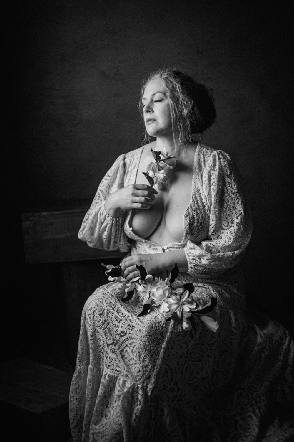 Black and white portrait of a woman in lace dress holding flowers, eyes closed, seated against dark background.