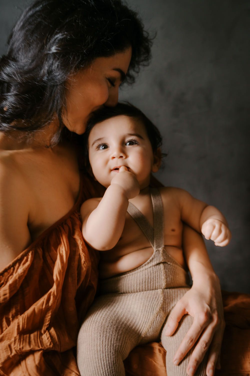 A mother tenderly kisses her baby's head while the infant sits in her arms, wearing a beige knit romper.