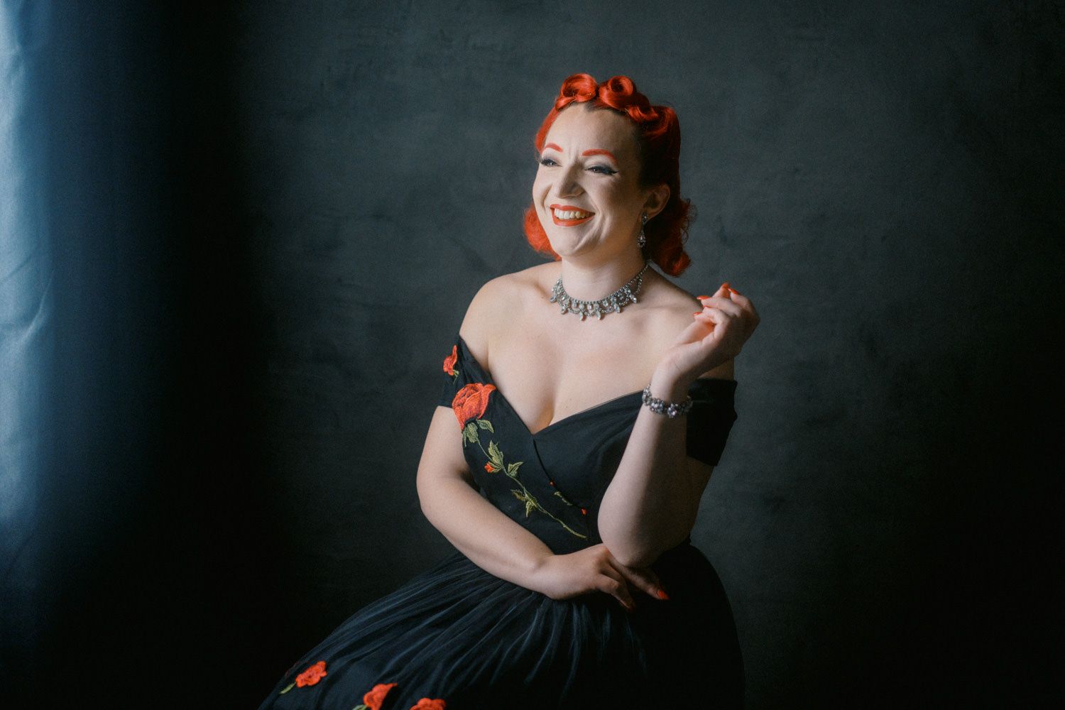 Woman in black off-shoulder gown with red floral accents and matching hair accessories, smiling elegantly in studio.
