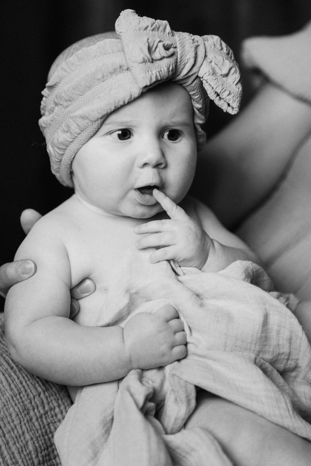 Black and white photo of an adorable baby wearing a bow headband, finger in mouth, wrapped in a soft blanket.