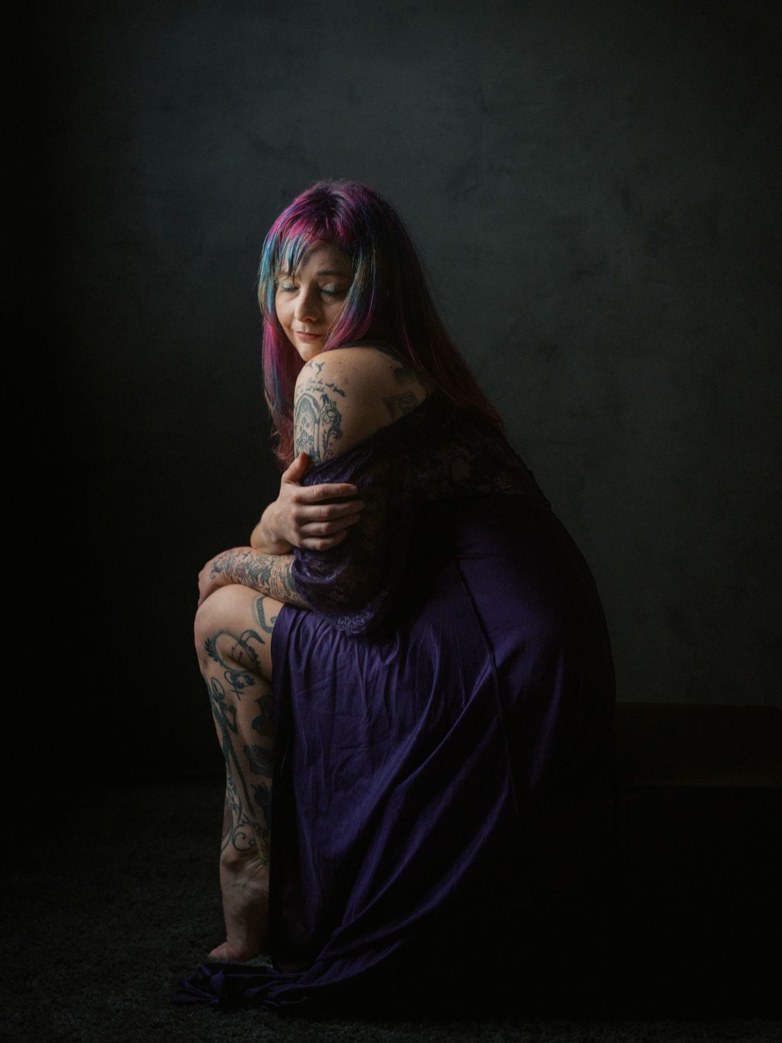 A tattooed woman with colorful hair sits in a purple dress, arms crossed, against a dark studio background.
