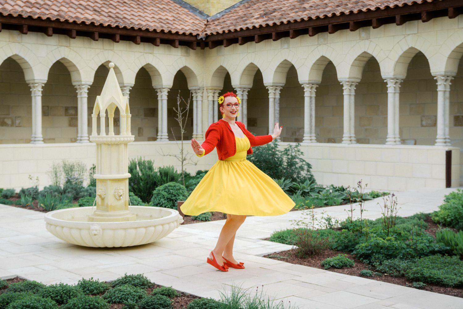 Woman in yellow skirt and red top twirling in a courtyard with arched columns and a fountain.