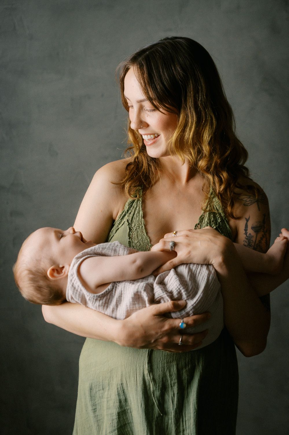 Smiling mother in green dress tenderly breastfeeding her newborn baby against a gray studio background.