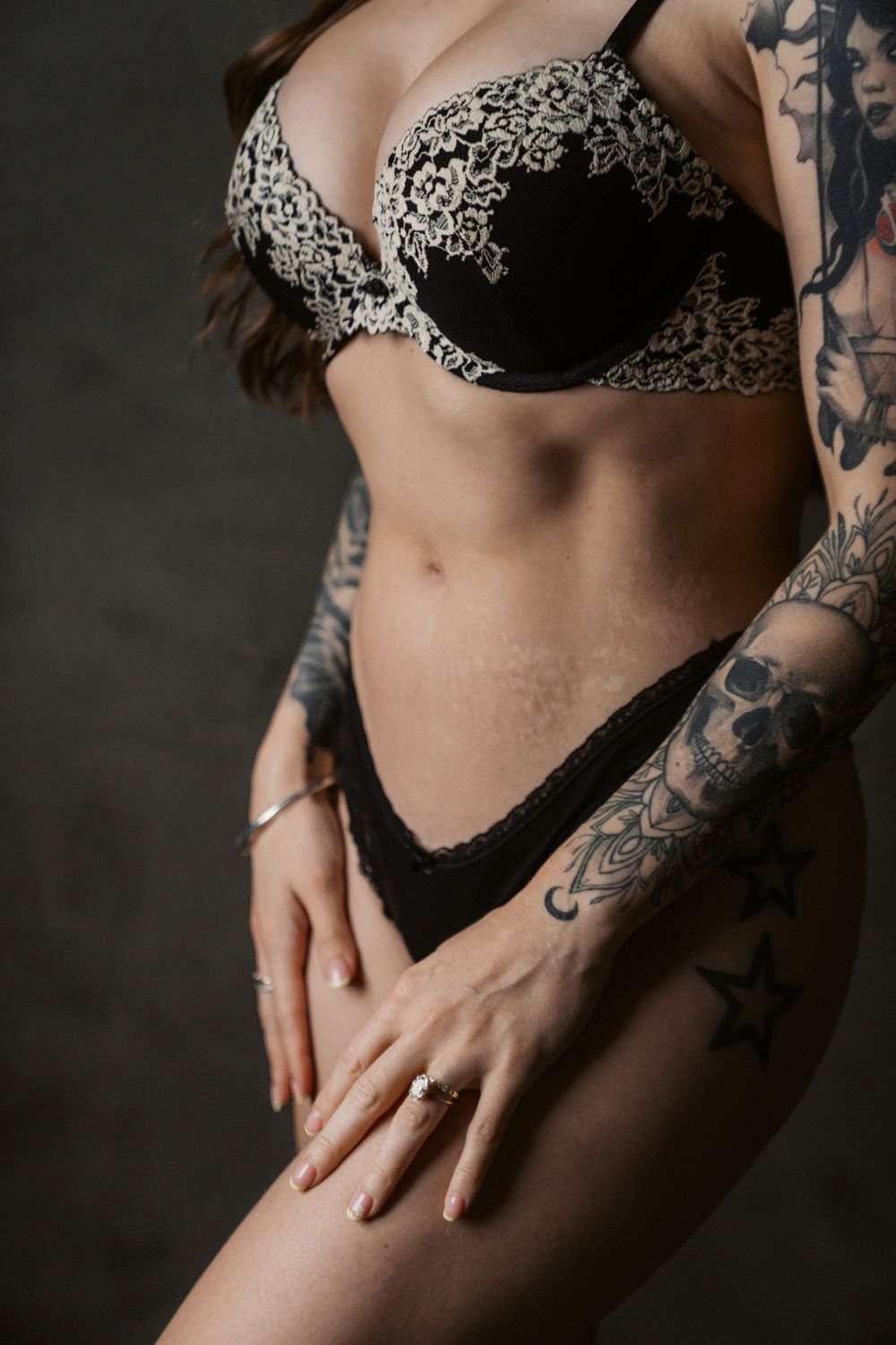 Woman in black lace lingerie set with tattoos on arms, posing against dark background.