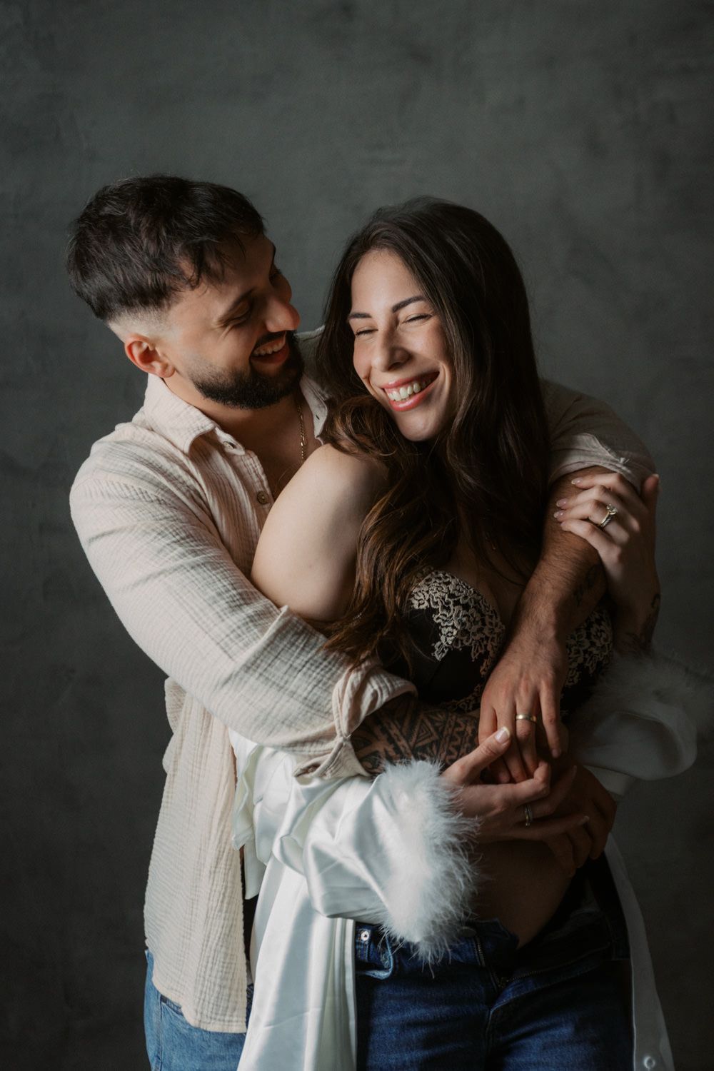 A smiling couple embraces warmly in a studio, the man hugging the laughing woman from behind against a gray background.
