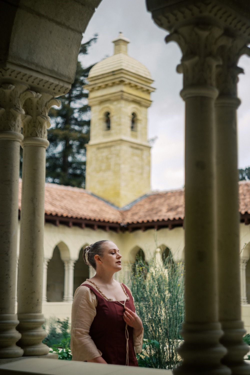 Woman in medieval dress stands among stone columns of a historic monastery with a bell tower backdrop.