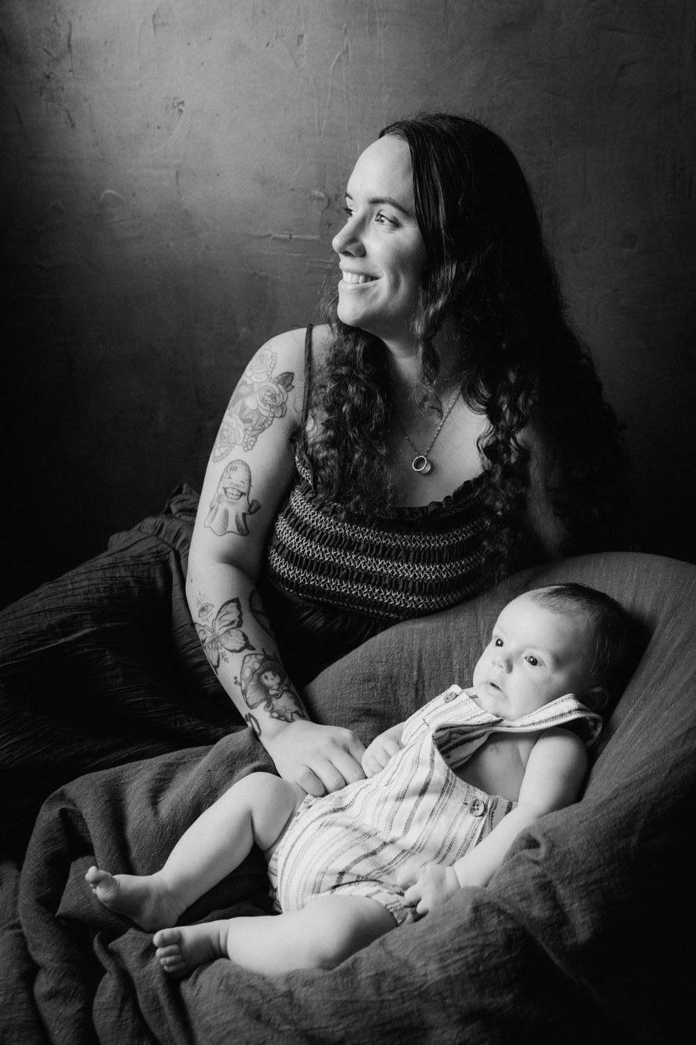 Black and white portrait of a smiling tattooed mother sitting with her infant baby in a studio setting.